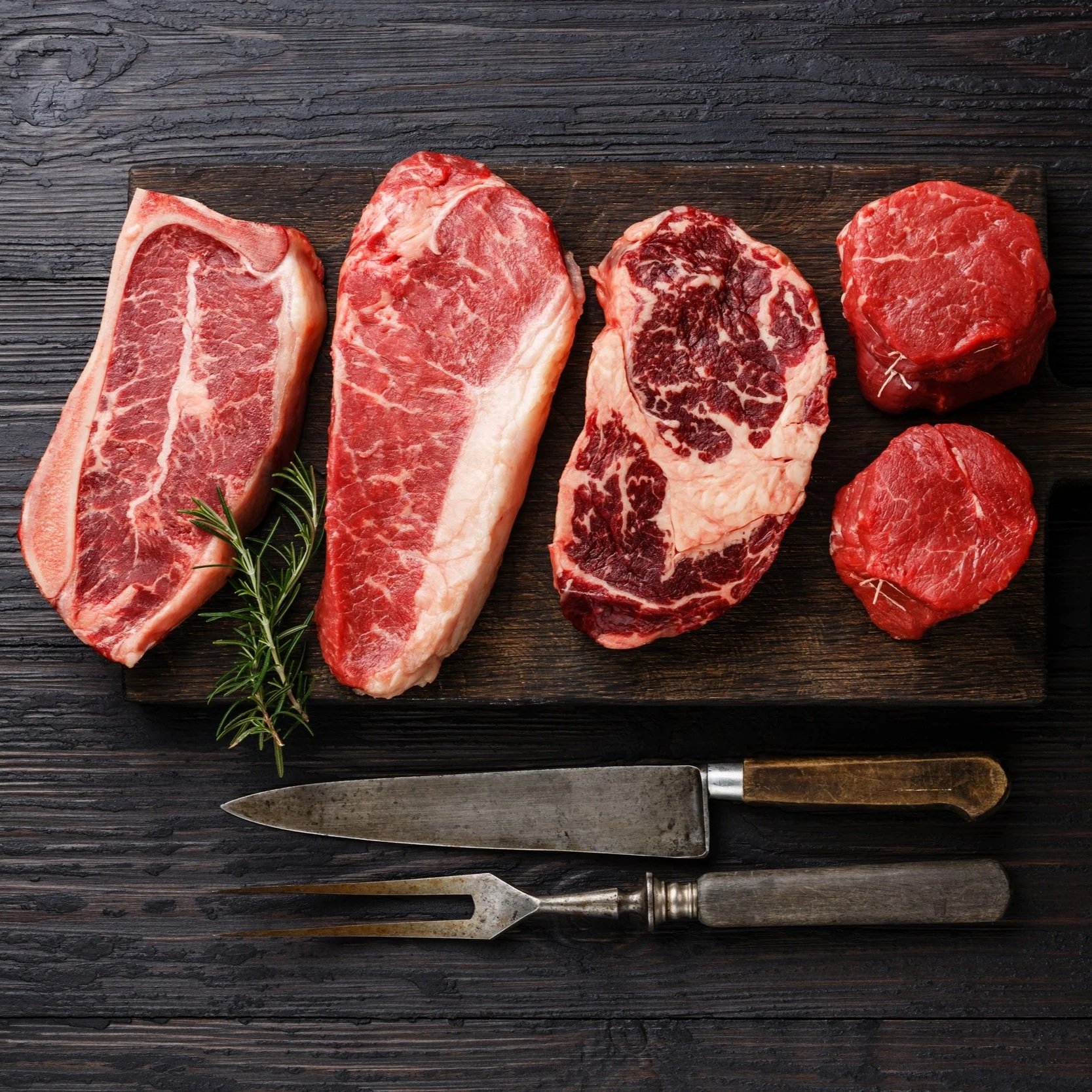 Beef_PremiumCuts_Bundle_Striploin_Ribeye_TenderloinFiletMignon Assorted cuts of raw beef, including ribeye, sirloin, and tenderloin, displayed on a wooden cutting board with a sprig of rosemary and vintage meat utensils. Ideal for cooking enthusiasts and butchery enthusiasts.